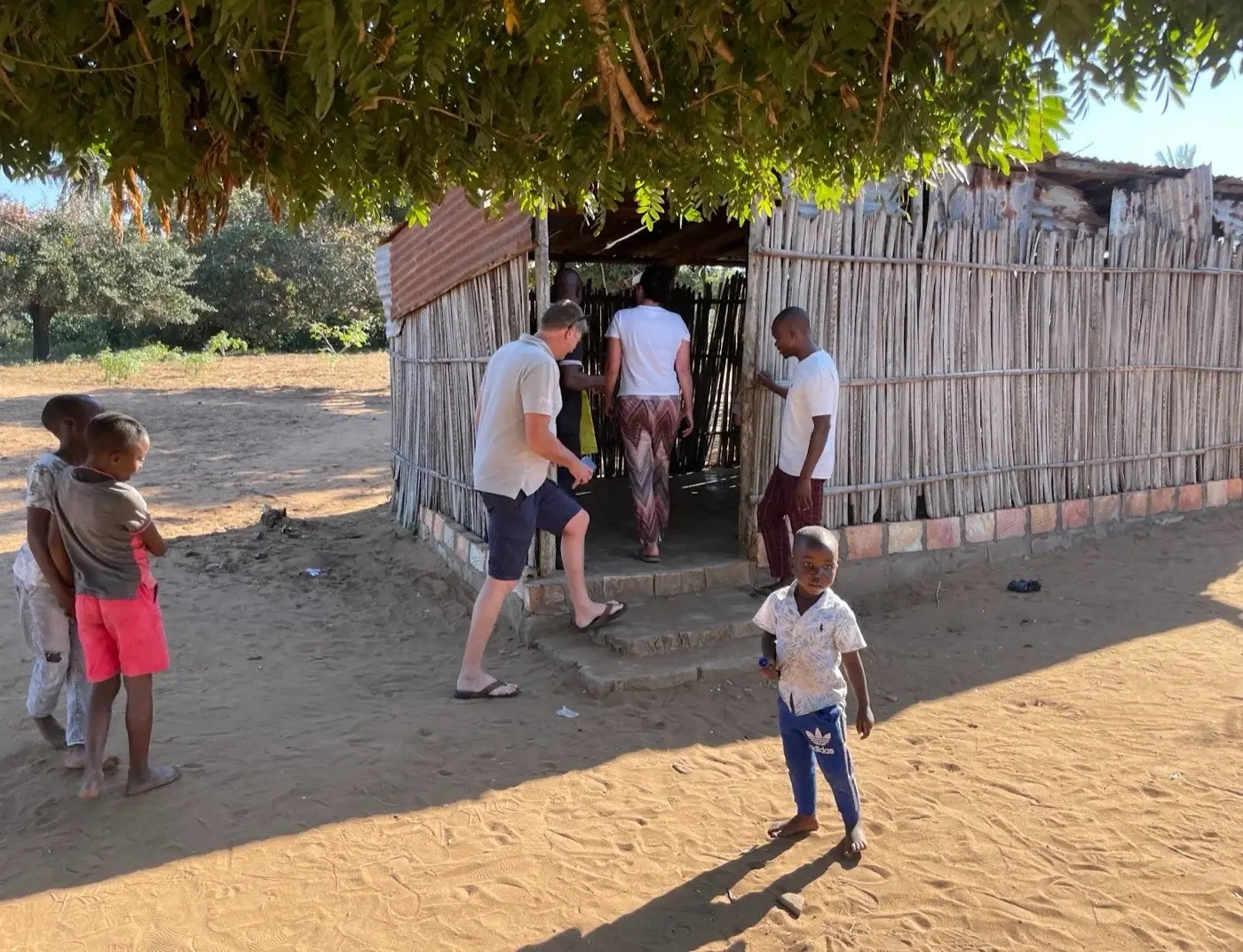 Außenansicht der Schulhütte aus Schilfrohr. Zwei Besucher gehen hinein, Kinder stehen im Sand davor.