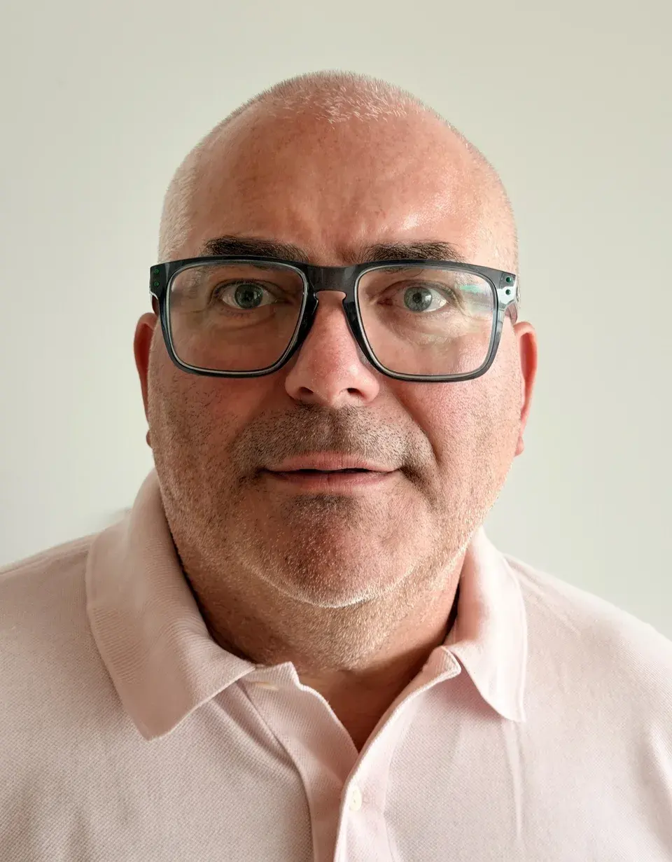 Dr. Alexander D. Boicev, Chair of the Board, with dark glasses and a light pink polo shirt, in front of a light-coloured wall