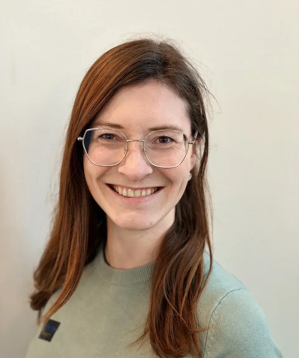 Natalie Bräunel, board member, with long auburn hair and glasses, smiling in front of a light-coloured wall
